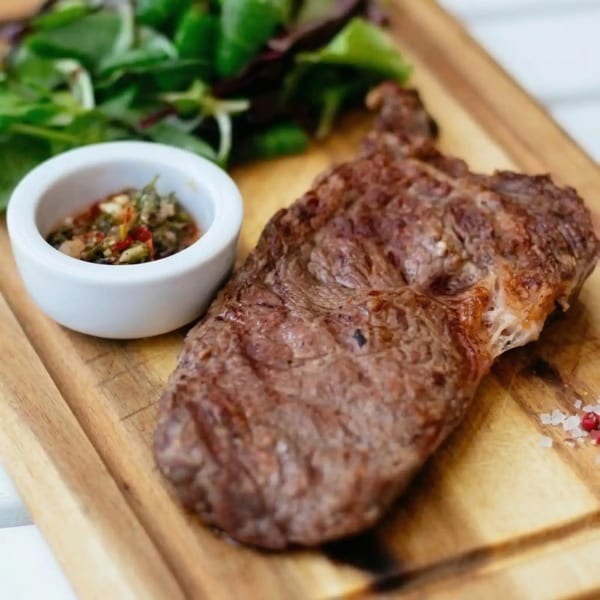 Grilled steak with a side of green salad and a small bowl of chimichurri sauce on a wooden board.